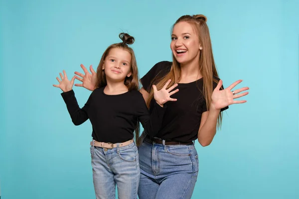 Mãe e filha com penteados engraçados, vestidos de camisas pretas e jeans jeans denim azul estão posando contra um fundo estúdio azul. Close-up shot. — Fotografia de Stock