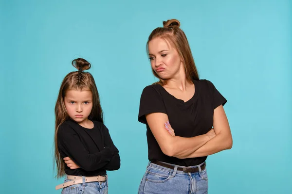 Mãe e filha com penteados engraçados, vestidos de camisas pretas e jeans jeans denim azul estão posando contra um fundo estúdio azul. Close-up shot. — Fotografia de Stock