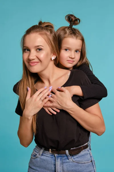 Mãe e filha com penteados engraçados, vestidos de camisas pretas e jeans jeans denim azul estão posando contra um fundo estúdio azul. Close-up shot. — Fotografia de Stock