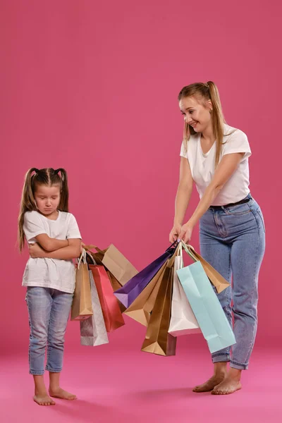 Mutter und Tochter mit Pferdeschwanz, gekleidet in weiße T-Shirts und blaue Jeans posieren vor rosa Hintergrund mit Paketen in der Hand. Volle Länge. — Stockfoto