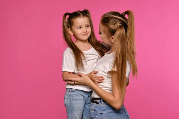 Mãe e filha com um rabo de cavalo engraçado, vestidas com t-shirts brancas e jeans denim azul estão posando contra um fundo estúdio rosa. Close-up shot. — Fotografia de Stock