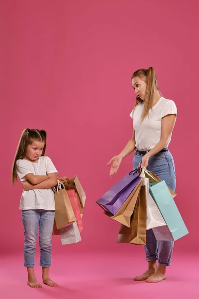 Mam en dochter met paardenstaarten, gekleed in witte t-shirts en blauwe jeans poseren tegen een roze achtergrond met pakjes in handen. Totale lengte. — Stockfoto