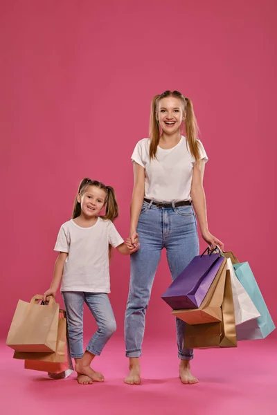 Mutter und Tochter mit Pferdeschwanz, gekleidet in weiße T-Shirts und blaue Jeans posieren vor rosa Hintergrund mit Paketen in der Hand. Volle Länge. — Stockfoto