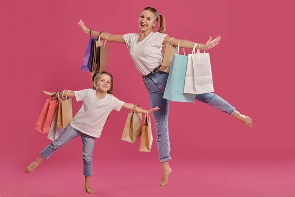 Maminka a dcera s culíky, oblečeni v bílých tričkách a modrých džínách, pózují na růžovém pozadí s balíčky v rukou. Plná délka. — Stock fotografie