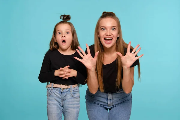 Mãe e filha com penteados engraçados, vestidos de camisas pretas e jeans jeans denim azul estão posando contra um fundo estúdio azul. Close-up shot. — Fotografia de Stock