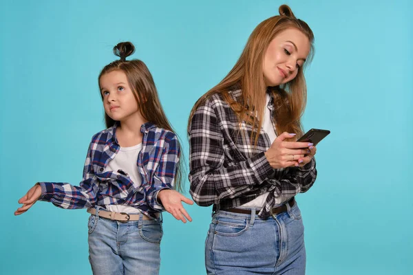 Mama i córka ubrane w szachowe koszule i niebieskie dżinsy jeansowe używają smartfona podczas pozowania na niebieskim tle studia. Strzał z bliska. — Zdjęcie stockowe