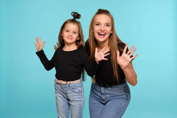 Mãe e filha com penteados engraçados, vestidos de camisas pretas e jeans jeans denim azul estão posando contra um fundo estúdio azul. Close-up shot. — Fotografia de Stock