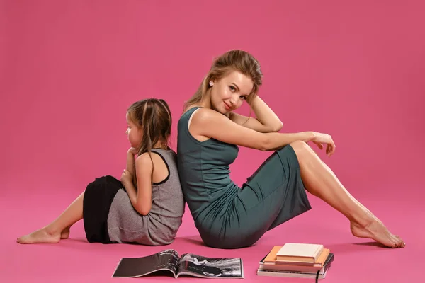 Anya és lánya szürke ruhákba öltözve ülnek a padlón magazinokkal és könyvekkel rózsaszín stúdió háttérrel. Közepes közelkép. — Stock Fotó