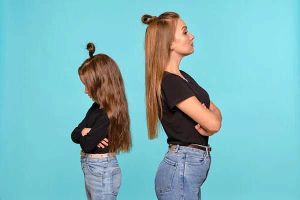 Mãe e filha com penteados engraçados, vestidos de camisas pretas e jeans jeans denim azul estão posando contra um fundo estúdio azul. Close-up shot. — Fotografia de Stock