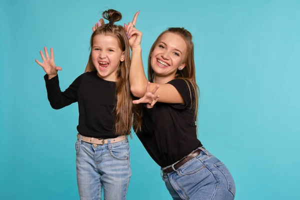 Mãe e filha com penteados engraçados, vestidos de camisas pretas e jeans jeans denim azul estão posando contra um fundo estúdio azul. Close-up shot. — Fotografia de Stock