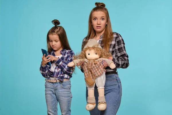 妈妈和女儿穿着格子衬衫和蓝色斜纹棉布牛仔裤，在蓝色工作室的背景下摆姿势时使用了智能手机。特写镜头. — 图库照片