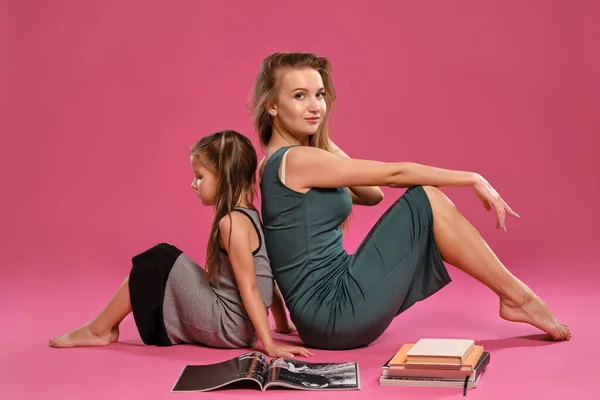 Mom and daughter dressed in gray dresses are posing sitting on a floor with magazine and books against pink studio background. Medium close-up shot.