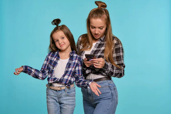 Mamma e figlia vestite con camicie a scacchi e jeans blu denim utilizzano smartphone mentre si pongono sullo sfondo di uno studio blu. Close-up shot. — Foto Stock