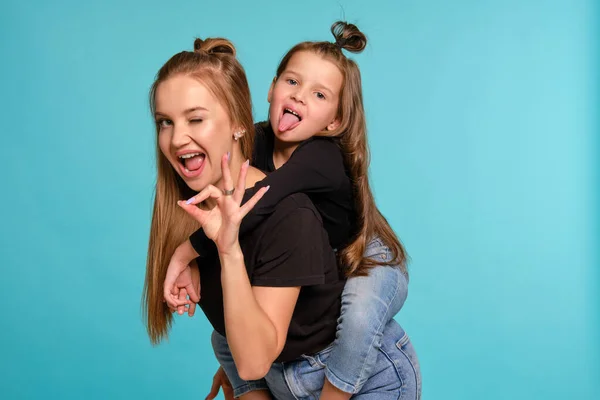 Mom and daughter with a funny hairstyles, dressed in black shirts and blue denim jeans are posing against a blue studio background. Close-up shot. — Stock Photo, Image