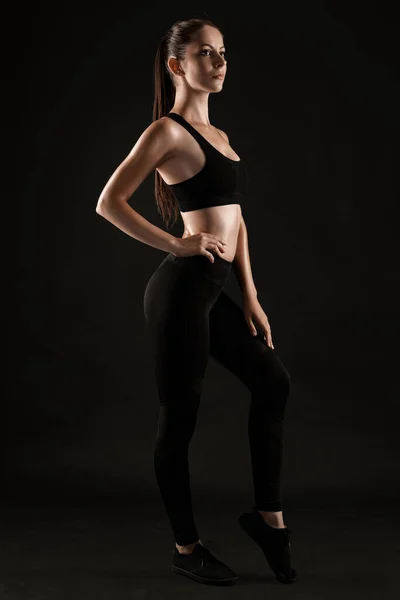 Brunette en jambières noires, haut et espadrilles se pose sur un fond noir. Condition physique, gymnase, concept de mode de vie sain. Longueur totale. — Photo