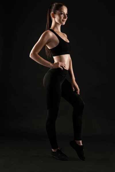 穿着黑色紧身裤、上衣和运动鞋的布鲁内特女人在黑色背景的衬托下摆姿势。健身，健身，健康的生活方式概念。全长. — 图库照片