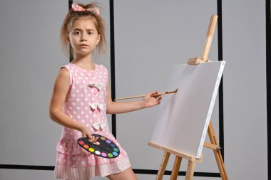 Pembe elbiseli küçük kız ressam resim sehpasının arkasında duruyor ve beyaz duvarlı bir sanat stüdyosunda fırçayla resim yapıyor. Orta boy yakın çekim..