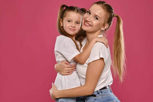 Mãe e filha com um rabo de cavalo engraçado, vestidas com t-shirts brancas e jeans denim azul estão posando contra um fundo estúdio rosa. Close-up shot. — Fotografia de Stock