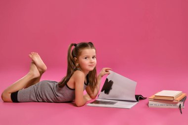 Gri elbiseli küçük bir kız, elinde dergi ve kitaplarla yerde pembe bir stüdyo arka planında poz veriyor. Orta boy yakın çekim..