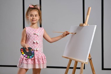 Pembe elbiseli küçük kız ressam resim sehpasının arkasında duruyor ve beyaz duvarlı bir sanat stüdyosunda fırçayla resim yapıyor. Orta boy yakın çekim..