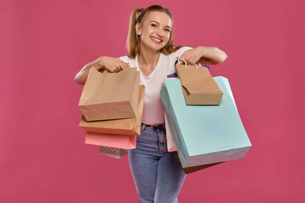 Blond vrouwtje met paardenstaarten, gekleed in wit t-shirt en jeans poserend tegen een roze achtergrond met pakjes. Een close-up. Oprechte emoties, winkelen. — Stockfoto