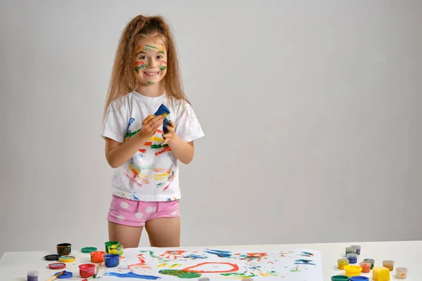 Little girl in white t-shirt standing at table with whatman and colorful paints, playing with a sponge soaked in paint. Isolated on white. Close-up.