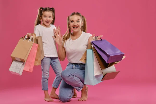 Mamá e hija con colas, vestida de camisetas blancas y vaqueros azules se ponen sobre fondo rosado con paquetes en las manos. Larga duración. — Foto de Stock