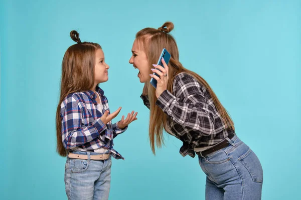 Мама и дочь, одетые в черные рубашки и синие джинсы, пользуются смартфоном, позируя на голубом студийном фоне. Закрытый снимок. — стоковое фото