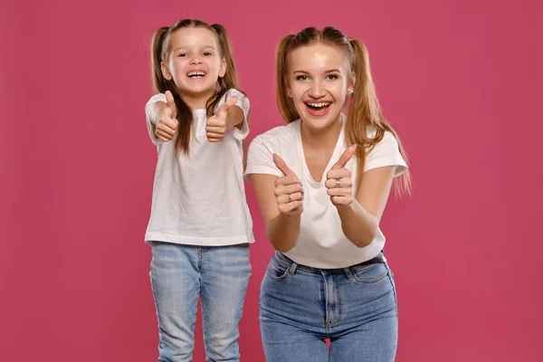 Mãe e filha com um rabo de cavalo engraçado, vestidas com t-shirts brancas e jeans denim azul estão posando contra um fundo estúdio rosa. Close-up shot. — Fotografia de Stock