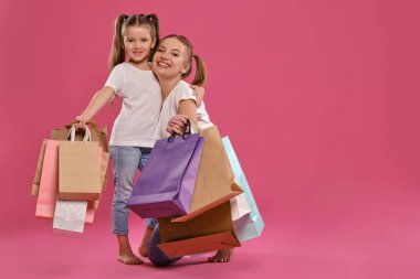 Beyaz tişört ve kot pantolon giymiş anne ve kız ellerinde paketlerle pembe arka planda poz veriyorlar. Tam uzunluk.