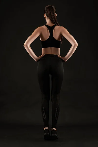 Brunette woman in black leggings, top and sneakers is posing against a black background. Fitness, gym, healthy lifestyle concept. Full length.