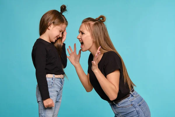 Mãe e filha com penteados engraçados, vestidos de camisas pretas e jeans jeans denim azul estão posando contra um fundo estúdio azul. Close-up shot. — Fotografia de Stock