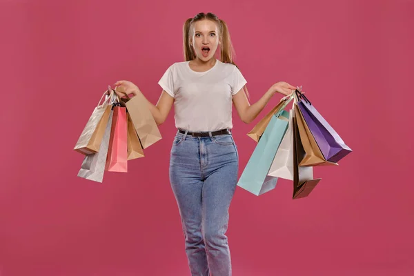 Blonde Frau mit Pferdeschwanz, in weißem T-Shirt und Jeans posiert sie vor rosa Hintergrund mit Paketen. Nahaufnahme. Aufrichtige Emotionen, Einkaufen. — Stockfoto
