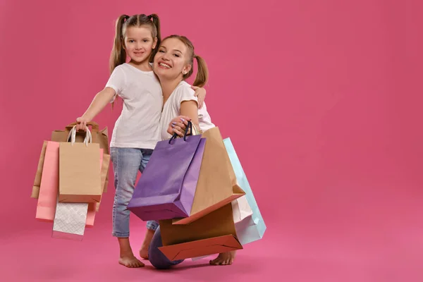 Mamá e hija con colas, vestida de camisetas blancas y vaqueros azules se ponen sobre fondo rosado con paquetes en las manos. Larga duración. — Foto de Stock