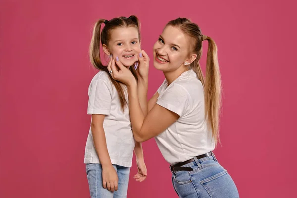 Mãe e filha com um rabo de cavalo engraçado, vestidas com t-shirts brancas e jeans denim azul estão posando contra um fundo estúdio rosa. Close-up shot. — Fotografia de Stock