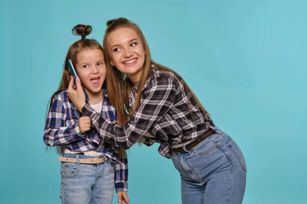 Mãe e filha vestidas com camisas de xadrez e jeans jeans jeans azuis estão usando smartphone enquanto posa contra um fundo estúdio azul. Close-up shot. — Fotografia de Stock