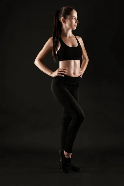 Mulher morena em leggings pretos, top e tênis está posando contra um fundo preto. Fitness, ginásio, conceito de estilo de vida saudável. Comprimento total. — Fotografia de Stock