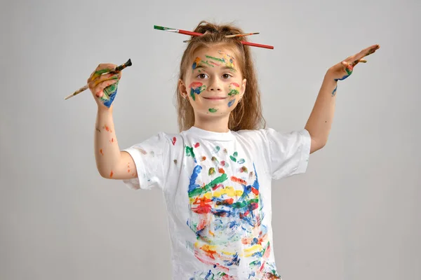 Little girl in white t-shirt, with brushes in her hair is posing standing isolated on white, gesticulating with painted hands and face. Close-up. — Stock Photo, Image