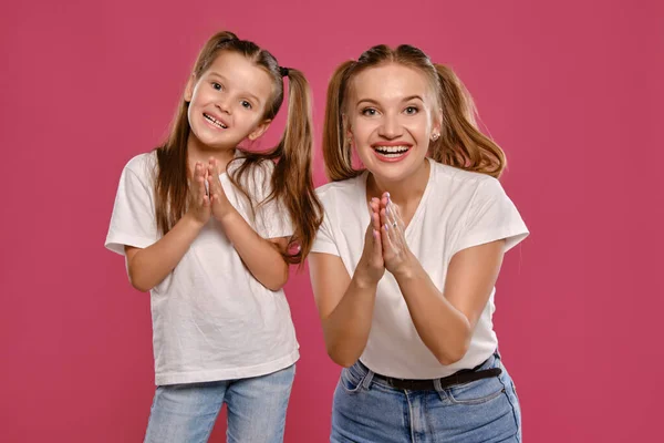 Mãe e filha com um rabo de cavalo engraçado, vestidas com t-shirts brancas e jeans denim azul estão posando contra um fundo estúdio rosa. Close-up shot. — Fotografia de Stock