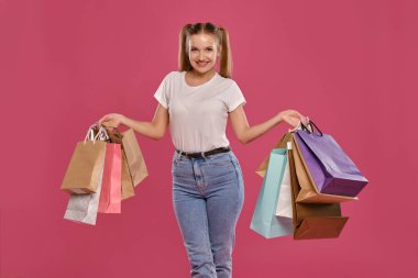 Atkuyruklu sarışın kadın, beyaz tişört ve kot pantolon giymiş pembe arka planda paketli poz veriyor. Yakın plan. Samimi duygular, alışveriş..