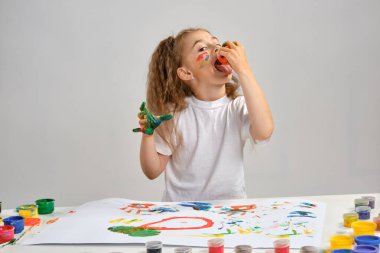 Beyaz tişörtlü küçük bir kız masasında hangi adamla oturuyormuş ve üzerine boyanmış yüz ve ellerle poz veriyormuş. Beyazda izole edilmiş. Orta boy yakın çekim.