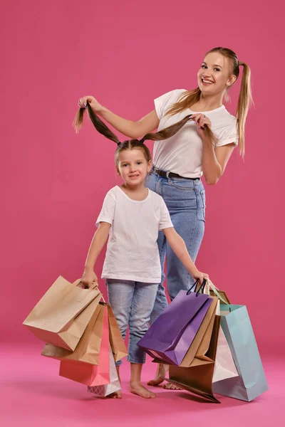 Mamá e hija con colas, vestida de camisetas blancas y vaqueros azules se ponen sobre fondo rosado con paquetes en las manos. Larga duración. — Foto de Stock