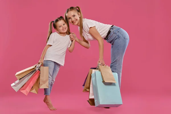 Mamá e hija con colas, vestida de camisetas blancas y vaqueros azules se ponen sobre fondo rosado con paquetes en las manos. Larga duración. — Foto de Stock