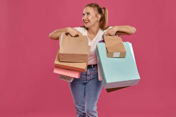 Blonde Frau mit Pferdeschwanz, in weißem T-Shirt und Jeans posiert sie vor rosa Hintergrund mit Paketen. Nahaufnahme. Aufrichtige Emotionen, Einkaufen. — Stockfoto