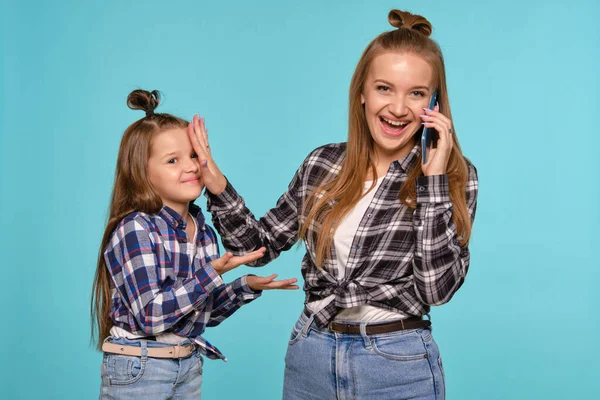 Mutter und Tochter in karierten Hemden und blauen Jeans nutzen das Smartphone, während sie vor blauem Studiohintergrund posieren. Nahaufnahme. — Stockfoto