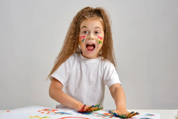 Klein meisje in wit t-shirt zittend aan tafel met whatman en verf erop, poserend met beschilderd gezicht en handen. Geïsoleerd op wit. Middelmatige close-up. — Stockfoto