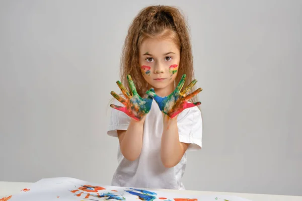 Little girl in white t-shirt sitting at table with whatman and paints on it, posing with painted face and hands. Isolated on white. Medium close-up. — Stock Photo, Image