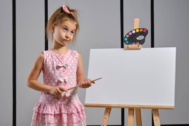 Pembe elbiseli küçük kız ressam resim sehpasının arkasında duruyor ve beyaz duvarlı bir sanat stüdyosunda fırçayla resim yapıyor. Orta boy yakın çekim..