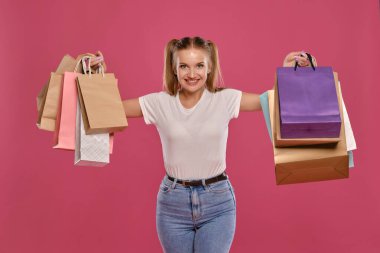 Atkuyruklu sarışın kadın, beyaz tişört ve kot pantolon giymiş pembe arka planda paketli poz veriyor. Yakın plan. Samimi duygular, alışveriş..