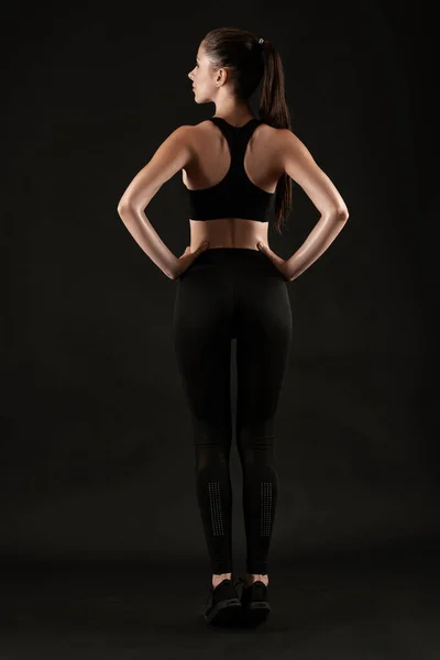 Mulher morena em leggings pretos, top e tênis está posando contra um fundo preto. Fitness, ginásio, conceito de estilo de vida saudável. Comprimento total. — Fotografia de Stock